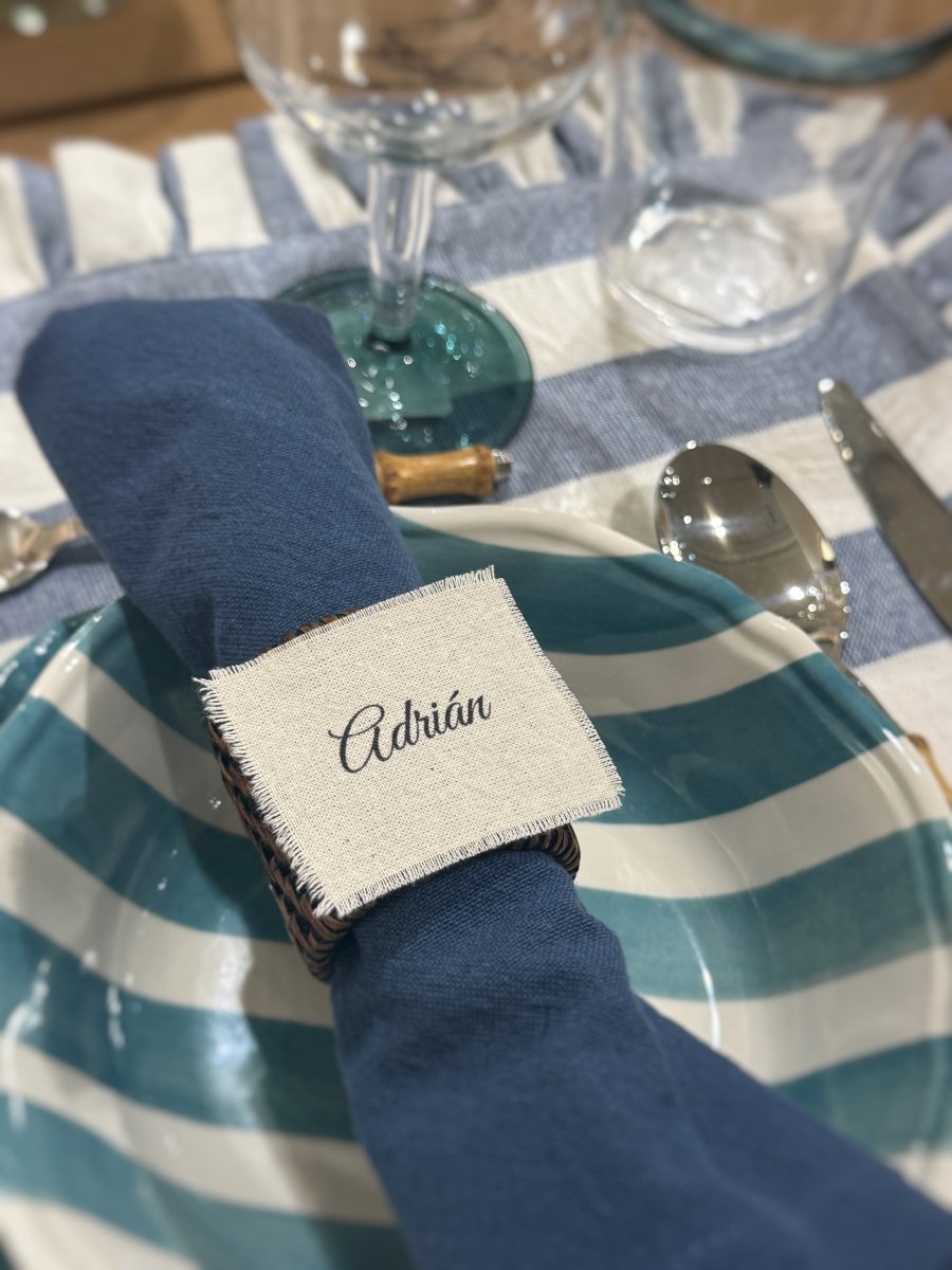 Marcasitios de algodón deshilachado personalizado con el nombre 'Adrián' en caligrafía elegante, presentado sobre una servilleta azul y un plato de rayas.