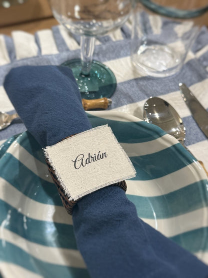Marcasitios de algodón deshilachado personalizado con el nombre 'Adrián' en caligrafía elegante, presentado sobre una servilleta azul y un plato de rayas.
