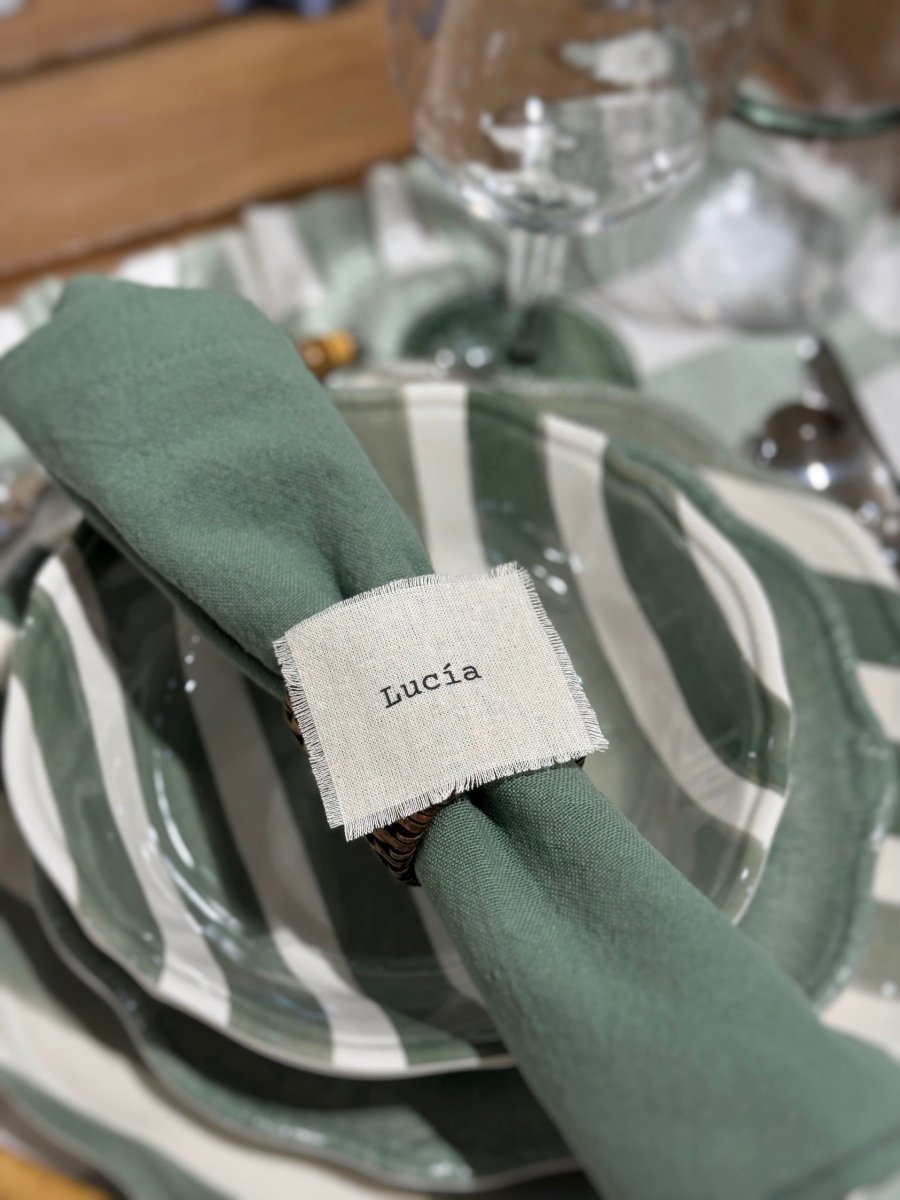 Marcasitios de algodón deshilachado personalizado con el nombre 'Lucía' sobre una servilleta de lino verde en una mesa de boda coordinada con vajilla de rayas.